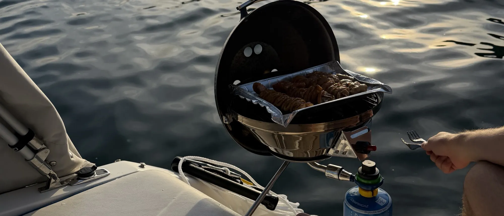 Kleiner Grill an Bord, dahinter das Wasser im Abendlicht.